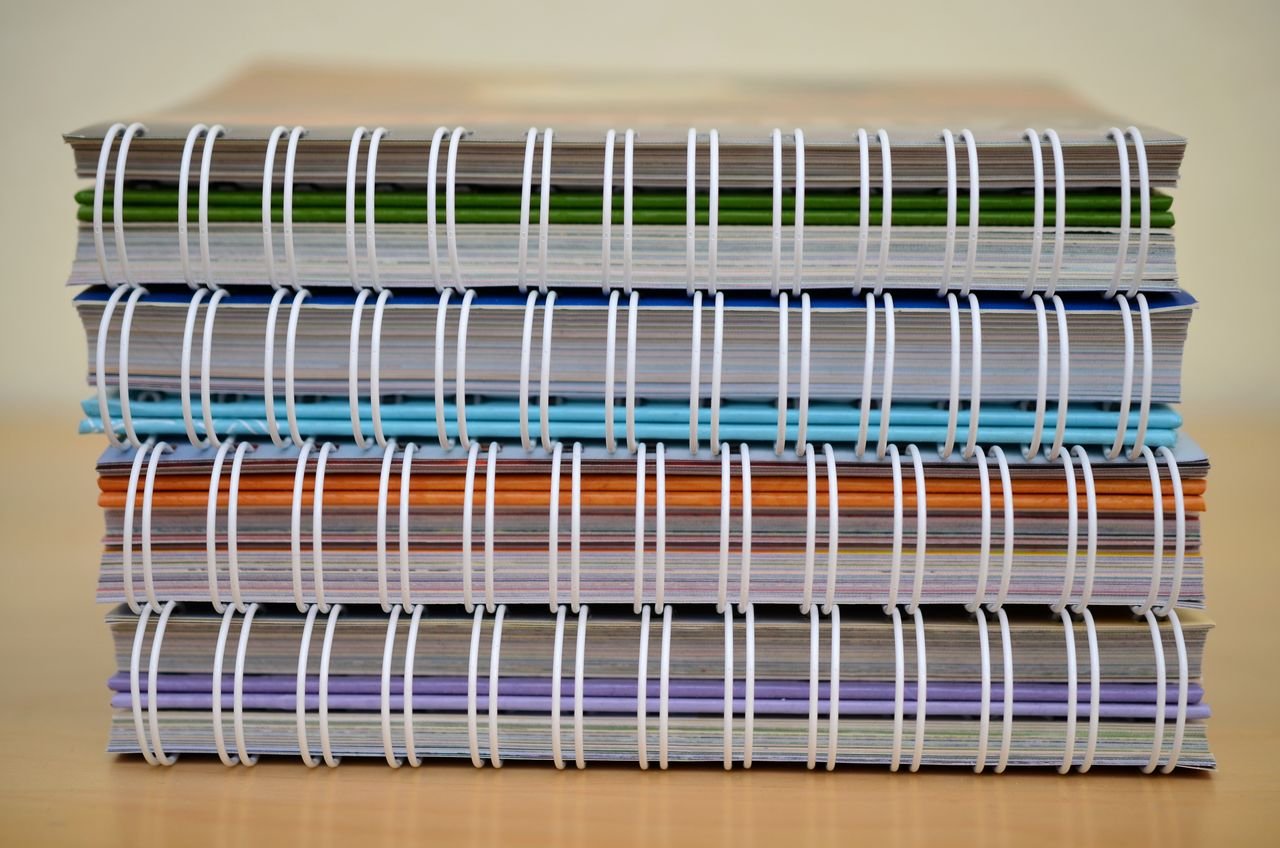 A stack of colorful spiral-bound notebooks placed on a wooden table, showcasing various colors and designs.