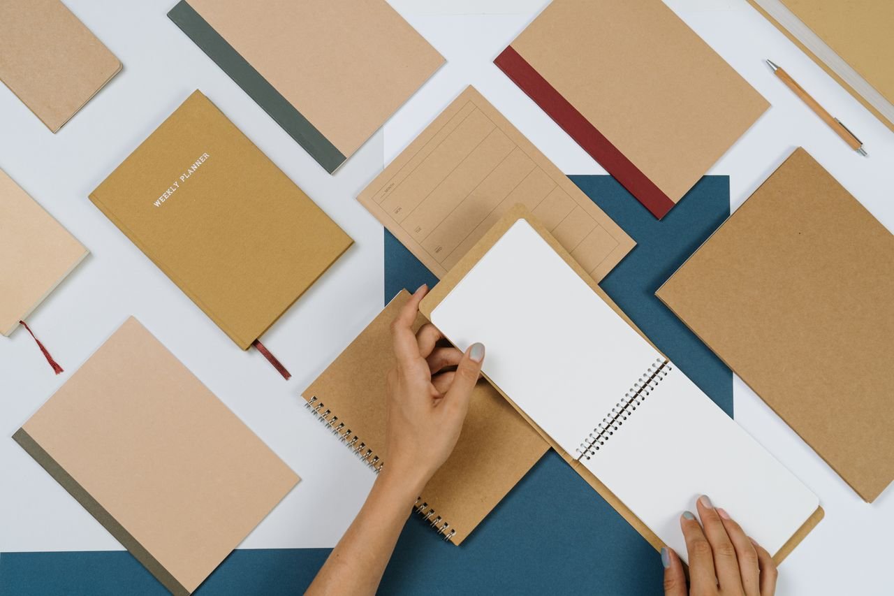Hands flipping a spiral notepad amid brown stationery in a flat lay setup.